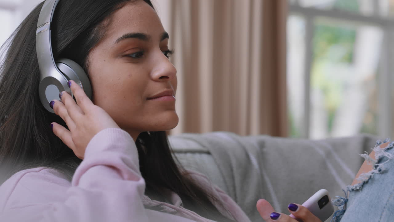 una adolescente feliz escuchando música con auriculares relajándose en el sofá en casa enviando mensajes de texto con su teléfono inteligente disfrutando de compartir su estilo de vida en las redes sociales