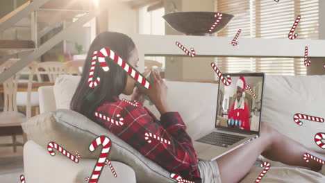 candy cane icons over woman drinking coffee and having a videocall on laptop during christmas