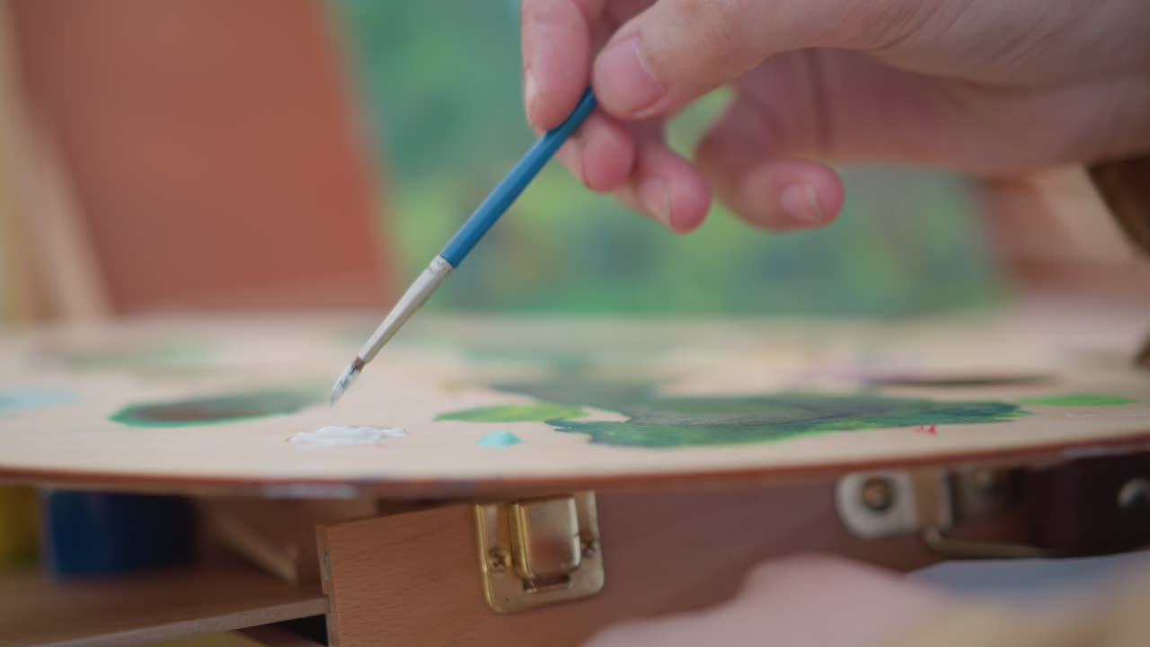 Artista concentrato che mescola colori su una palette di legno. Scena rilassante di un artista che unisce vivaci pigmenti verdi e blu. Artista calmo che combina attentamente le tonalità sulla palette in una tranquilla atmosfera da giardino