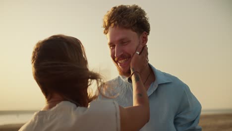 A-bearded-man-with-curly-hair-in-a-blue-shirt-is-talking-to-his-blonde-girlfriend,-who-is-stroking-his-chin-and-beard-against-the-yellow-sky-near-the-river