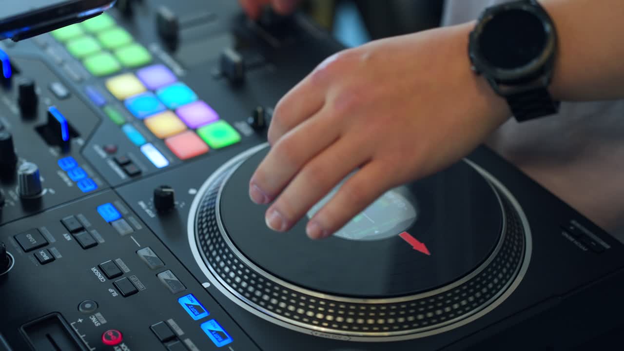 Primo piano in tempo reale della mano di un DJ che esegue scratching sulla jog wheel rotante del controller giradischi, pad di performance illuminati e colorati, pulsanti LED blu, fader e manopole visibili durante un live set