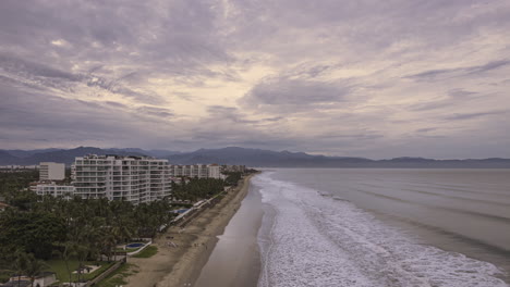 Hyperlapse-Aus-Der-Luft-Vom-Strand-Von-Puerto-Vallarta,-Hotels-Und-Bewölktem-Himmel,-Aufgenommen-Von-Einer-Vorwärtsfliegenden-Drohne,-Mit-Meereswellen-Und-Hügeln-Im-Hintergrund