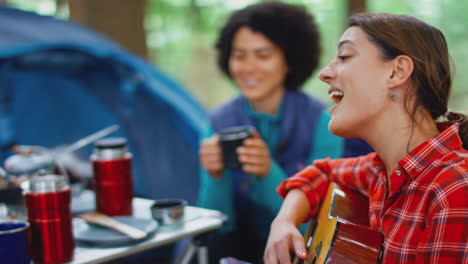 Group-Of-Female-Friends-On-Camping-Holiday-In-Forest-Eating-Meal-And-Singing-Along-To-Guitar