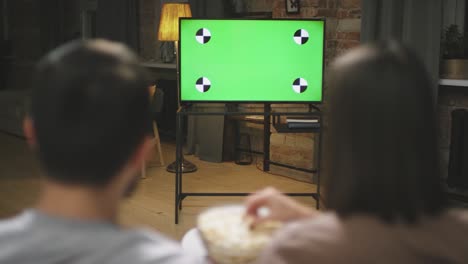 rear view of a couple sitting on the couch looking at a screen while eating popcorn