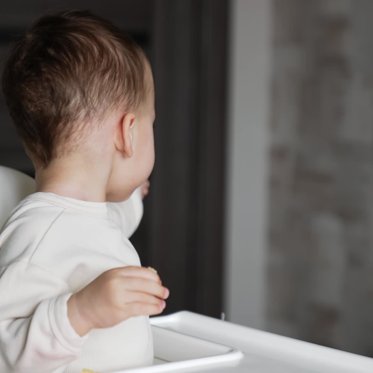 Caucasian kid sitting in feeding chair looking back. Child eats something pointing somewhere with his hand. Blurred backdrop