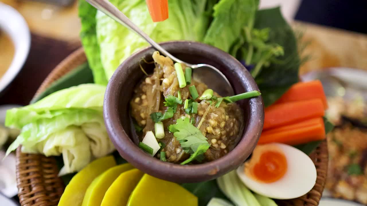 Hand dips assorted fresh vegetables into a traditional roasted green chili dip, served in a clay bowl with vibrant local produce under natural daylight