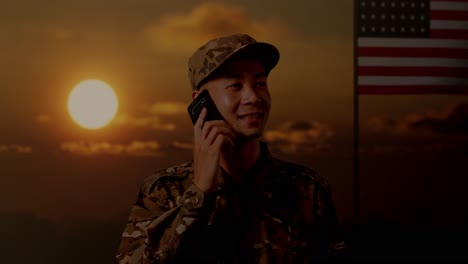 close up of asian man soldier talking on smartphone and looking around while standing with flag of the united states, sunset time