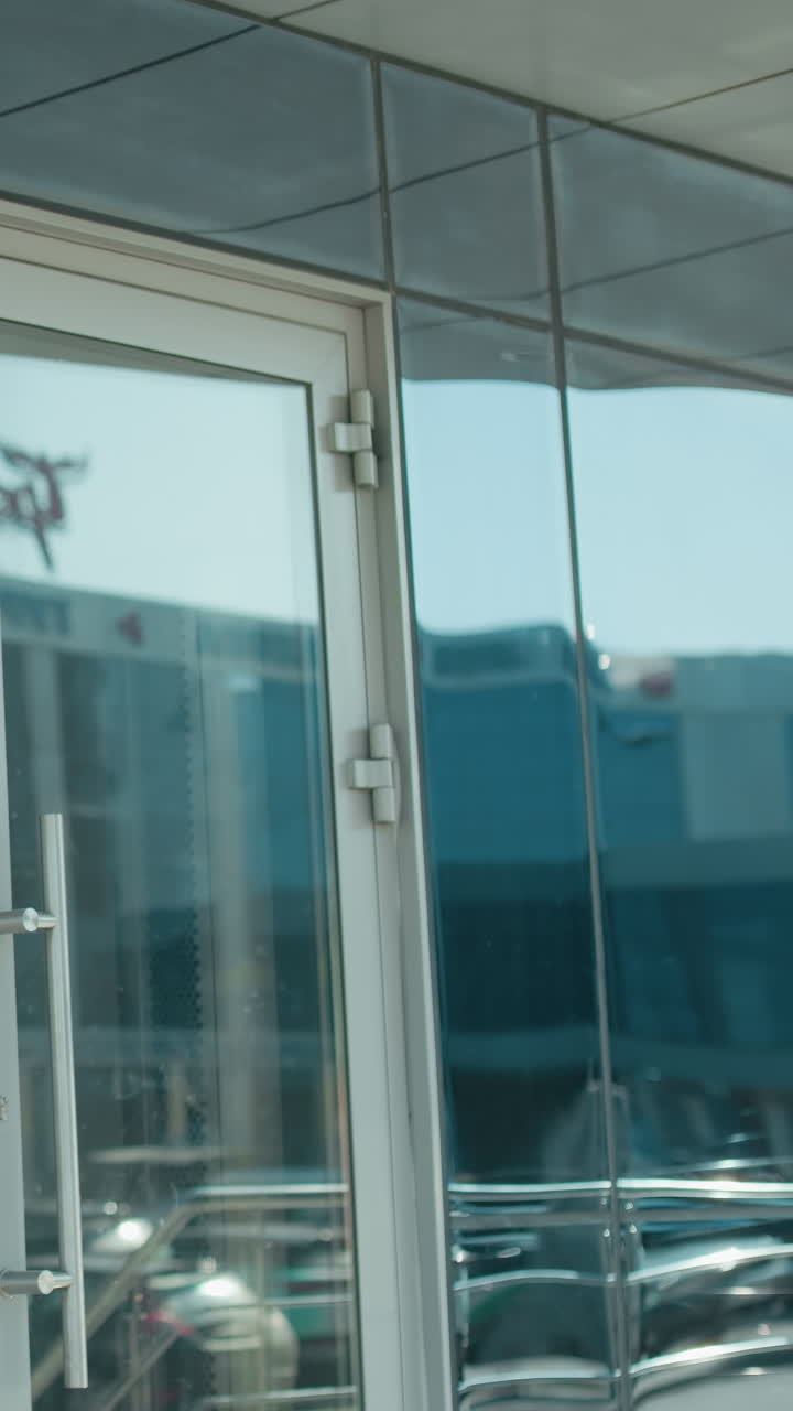 Business lady carrying handbag walks into office building dressed professionally in black jacket and gray pants, with glass door reflecting urban street, trees and parked cars