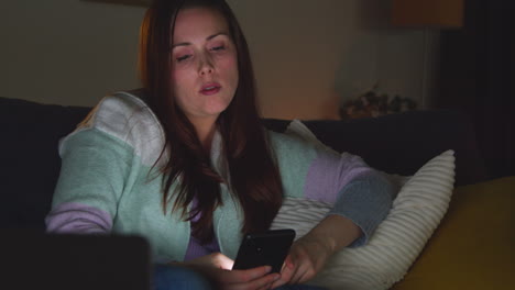 woman sitting on sofa at home at night streaming or watching movie or show on laptop and scrolling internet on phone 2