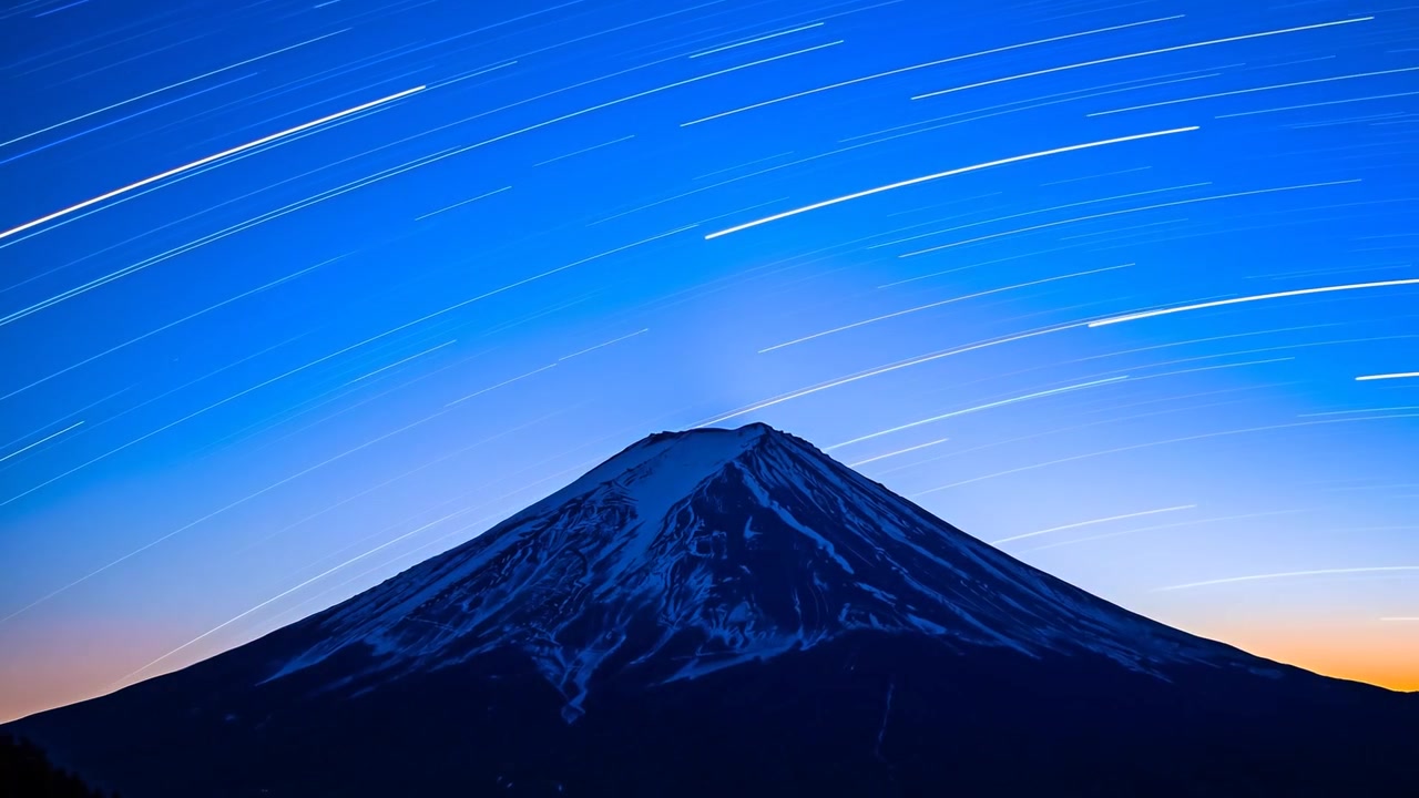タイマーを使って撮影中のカメラが、円錐形の雪山の上空に星の軌跡を捉え、地平線の輝きを見せている。