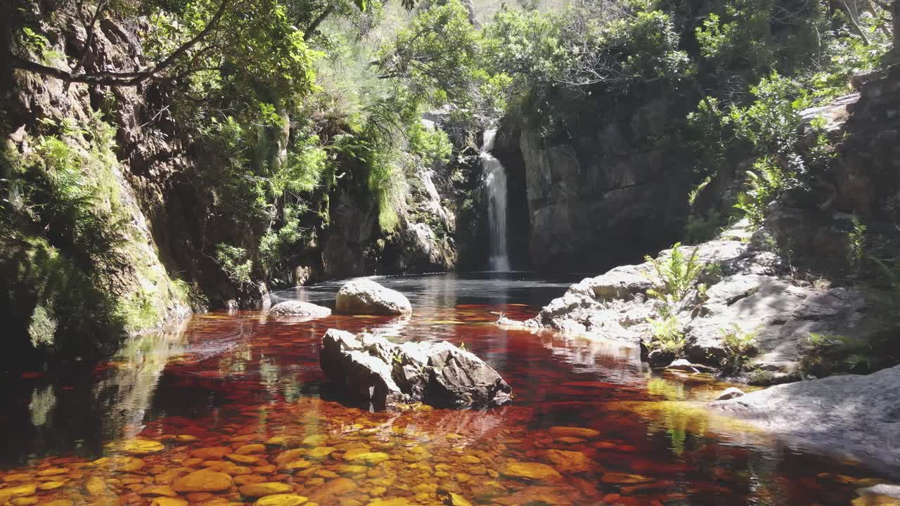 fluxo de água de aparência marrom e vermelha com cachoeira ao fundo e arredores com aparência de selva