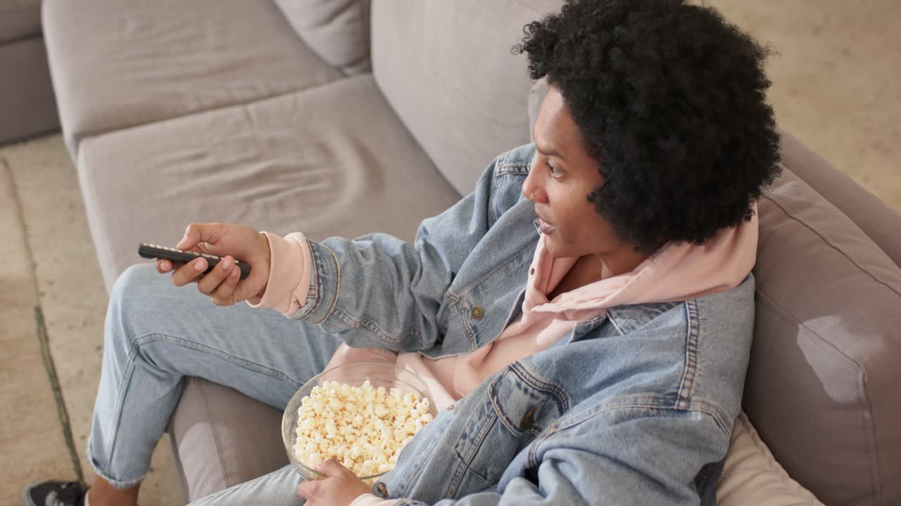 Afroamerikansk mand, der klikker med fjernbetjeningen og spiser popcorn i en grå sofa i stuen, kopiområde