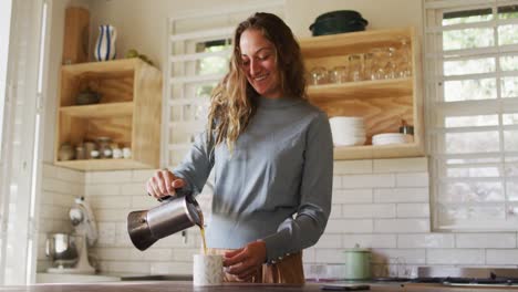 Happy-caucasian-woman-standing-in-cottage-kitchen-pouring-coffee-from-pot-and-smiling