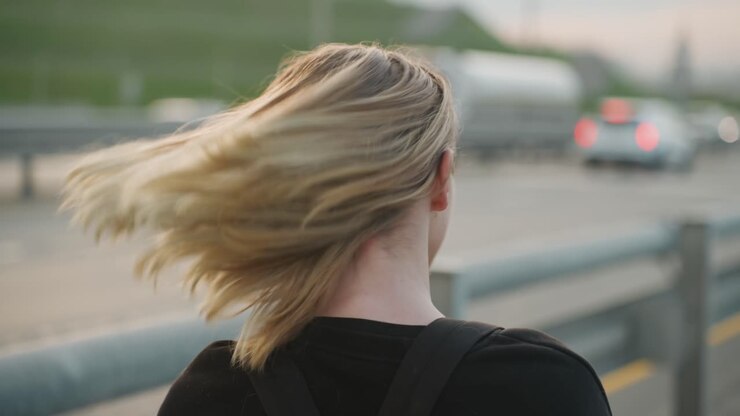 Giovane donna sulla strada che si sistema i capelli al crepuscolo, appoggiata a un guardrail accanto a un'autostrada trafficata con auto sfocate, coda di cavallo bionda sciolta al vento, luce soffusa del tramonto, felpa con cappuccio e zaino casual, contemplativa.
