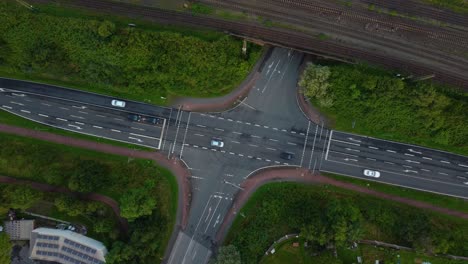 top-down-view-of-a-road-heading-all-directions-black-and-white-cars-driving-across-turning-left-and-right-trees-and-grass-along-the-roads-brown-train-tracks-at-the-top-of-the-frame-travel-work-feel