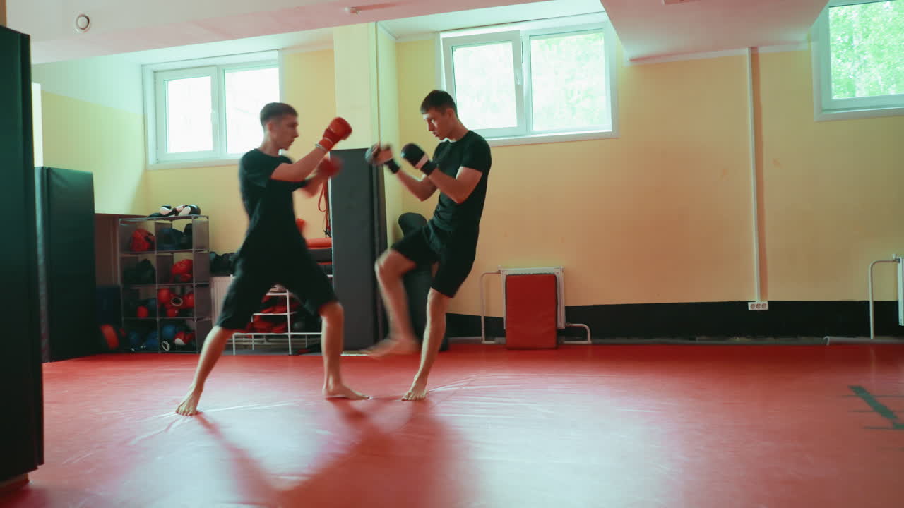 Two men practicing martial arts sparring on red mat in gym, training combat skills, wearing black sportswear, using boxing gloves and pads, focused on fighting technique under natural light from windows