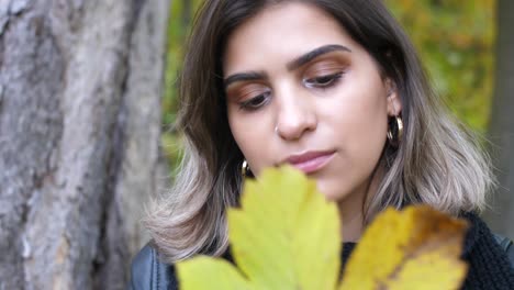 brunette girl in the autumn forest holds a yellow leaf in her hand and hides her face behind it - close shot