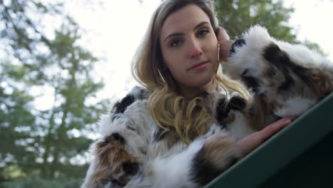 stylish young blonde woman wearing a fur jacket posing on a railing, forest background