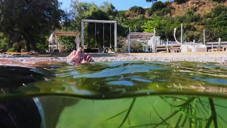 half underwater view of male legs and feet relaxing while floating on sea water with algae and event setup with swing on beach in background