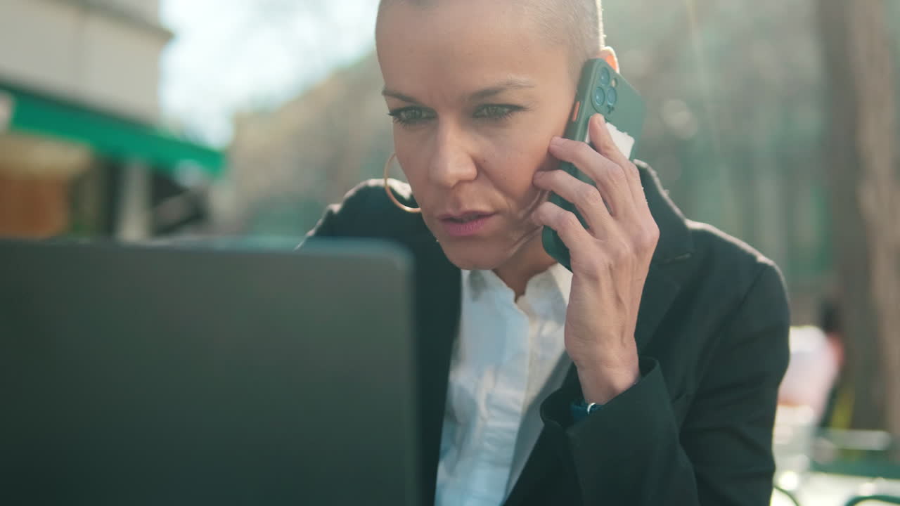 Focused Woman Talking on Phone While Working on Laptop Outdoors