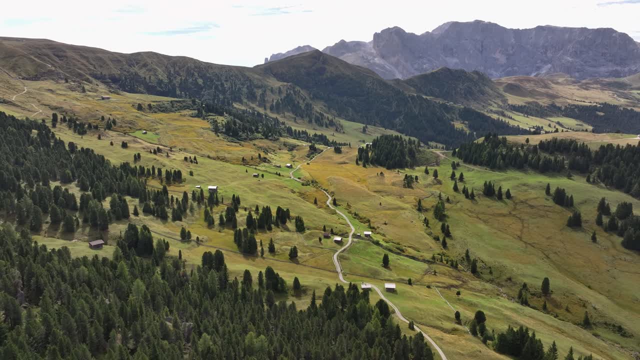Alpine meadows in the Dolomites from a bird's-eye view. A path winds through the meadows. Small houses are scattered across the meadows