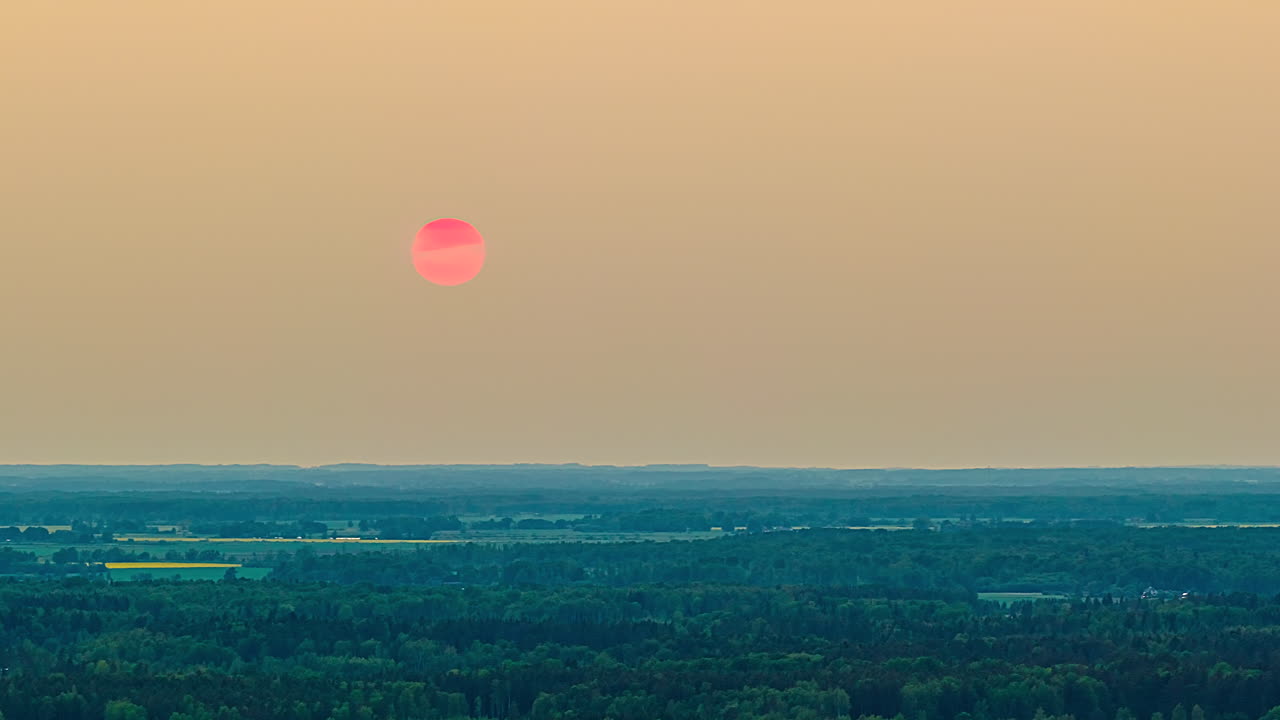 Drone hyperlapse of bright pink sun setting behind forest horizon under warm hazy sky