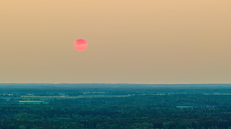 Drone hyperlapse of bright pink sun setting behind forest horizon under warm hazy sky