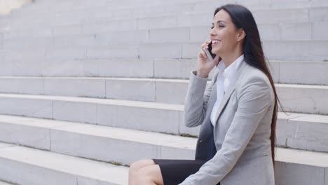 Lachende-Frau-Sitzt-Auf-Der-Treppe-Mit-Telefon