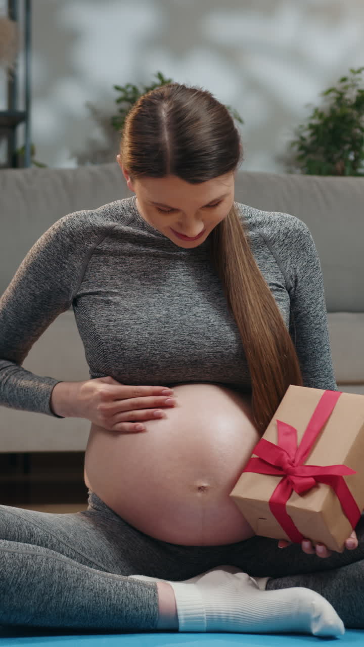 Expecting mother sits crosslegged indoors cradling her belly and holding a gift box with a red