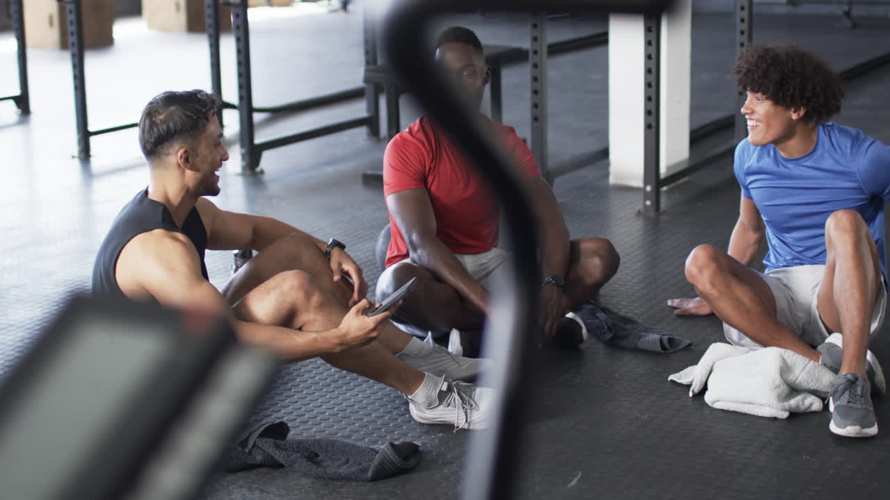 Group of diverse young men sitting on gym floor, talking and smiling