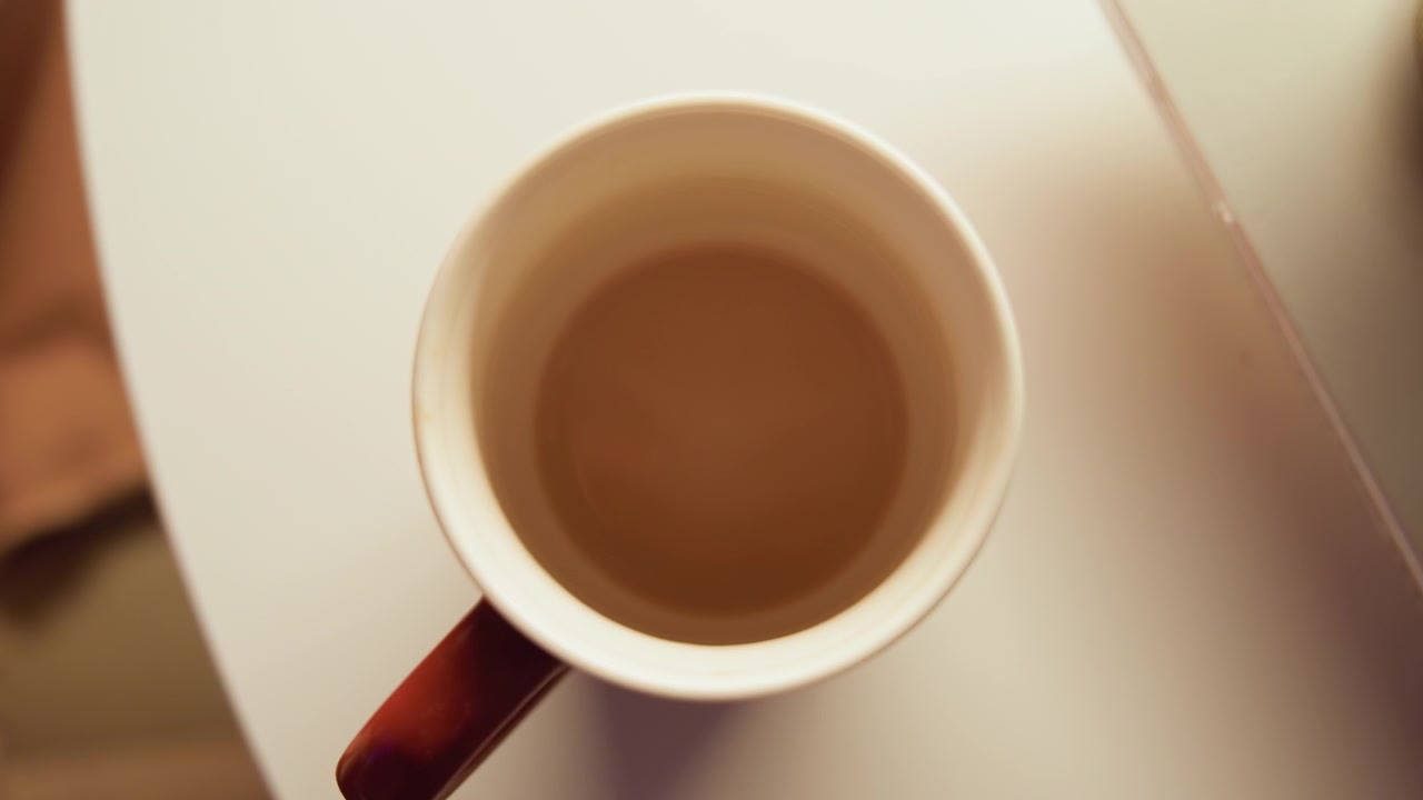 Una vista dall'alto mostra una tazza in ceramica con manico marrone parzialmente riempita con una bevanda marrone chiaro, mentre una mano si allunga per sollevare la tazza da un tavolo rotondo bianco, creando un movimento calmo in una luce interna accogliente.