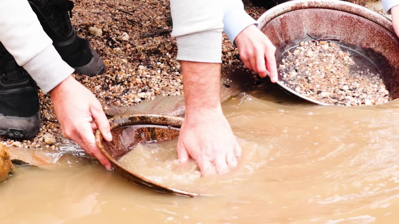 Close-up of hands using pans to sift through muddy water and gravel in search of gold.