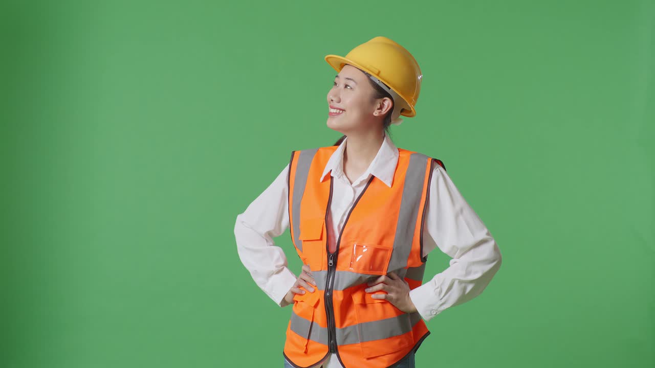 Asian Female Engineer Wearing Safety Helmet Looking Around While Standing With Arms Akimbo In The Green Screen Background Studio