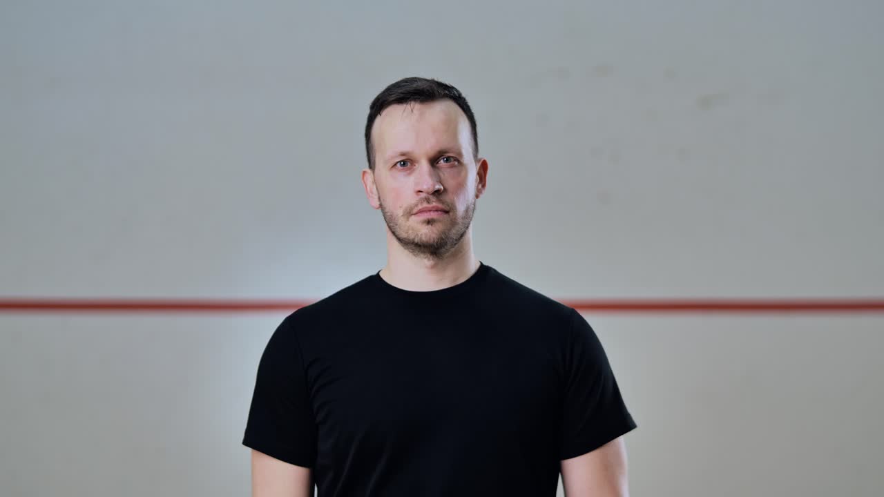 Man smiling and raising squash racket into lens focus in soft lit sports court