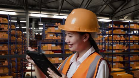 close up side view of asian female engineer with safety helmet standing in the warehouse with shelves full of delivery goods. taking note on the tablet and looking around in the storage