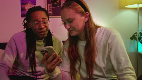 a young couple laughing together while looking at a phone in bed