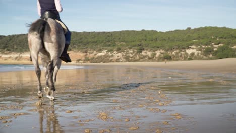 Tiro-De-Mano-Ascendente-En-Cámara-Lenta-En-Una-Hermosa-Playa-De-Arena-Durante-Olas-Tranquilas-Mientras-Dos-Jinetes-Galopan-Con-Sus-Caballos-Hacia-La-Libertad