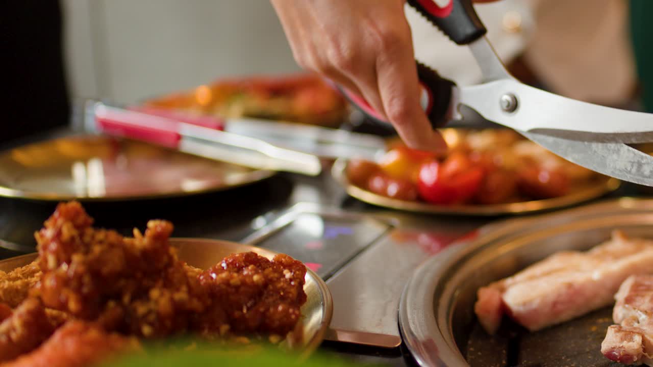 Person slices grilled meat on tabletop grill using scissors and tongs, warm indoor lighting, close-up