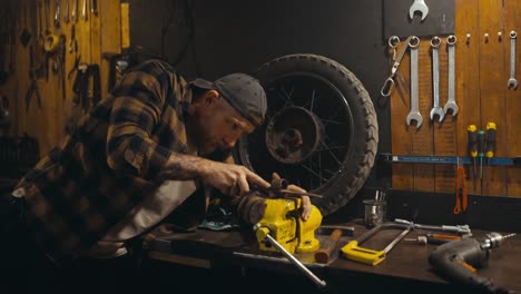 Confident-guy-mechanic-in-a-cap-and-plaid-shirt-works-with-a-file-on-a-clamp-on-a-workbench-in-his-workshop