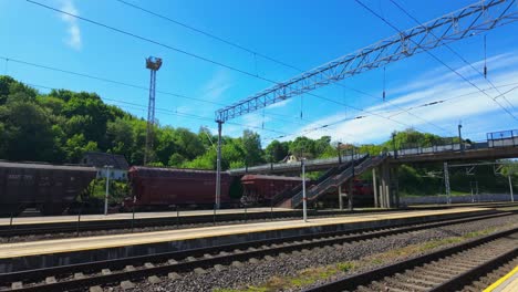 a quiet train station with empty platforms and rail tracks on a sunny day, featuring a bridge, power lines, and green trees in the background