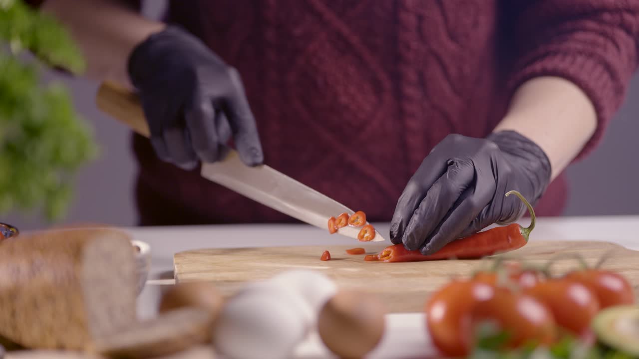 Slowmotion closeup of a chef slicing a red chili pepper on a wooden board. The sharp knife cuts smoothly, revealing fresh texture and seeds. Black gloves ensure hygiene, with tomatoes and herbs nearby