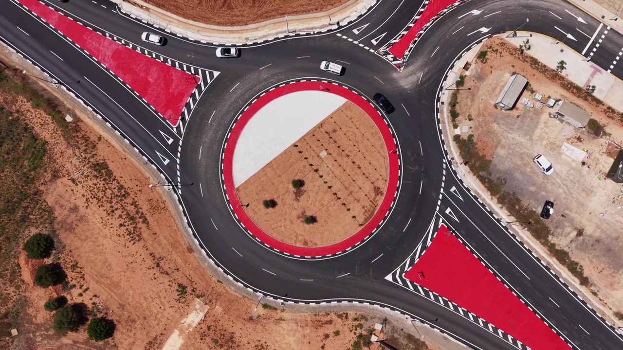 Overhead view of a large circular roundabout with red painted zones and white lane markings, several cars navigating the intersection surrounded by arid terrain