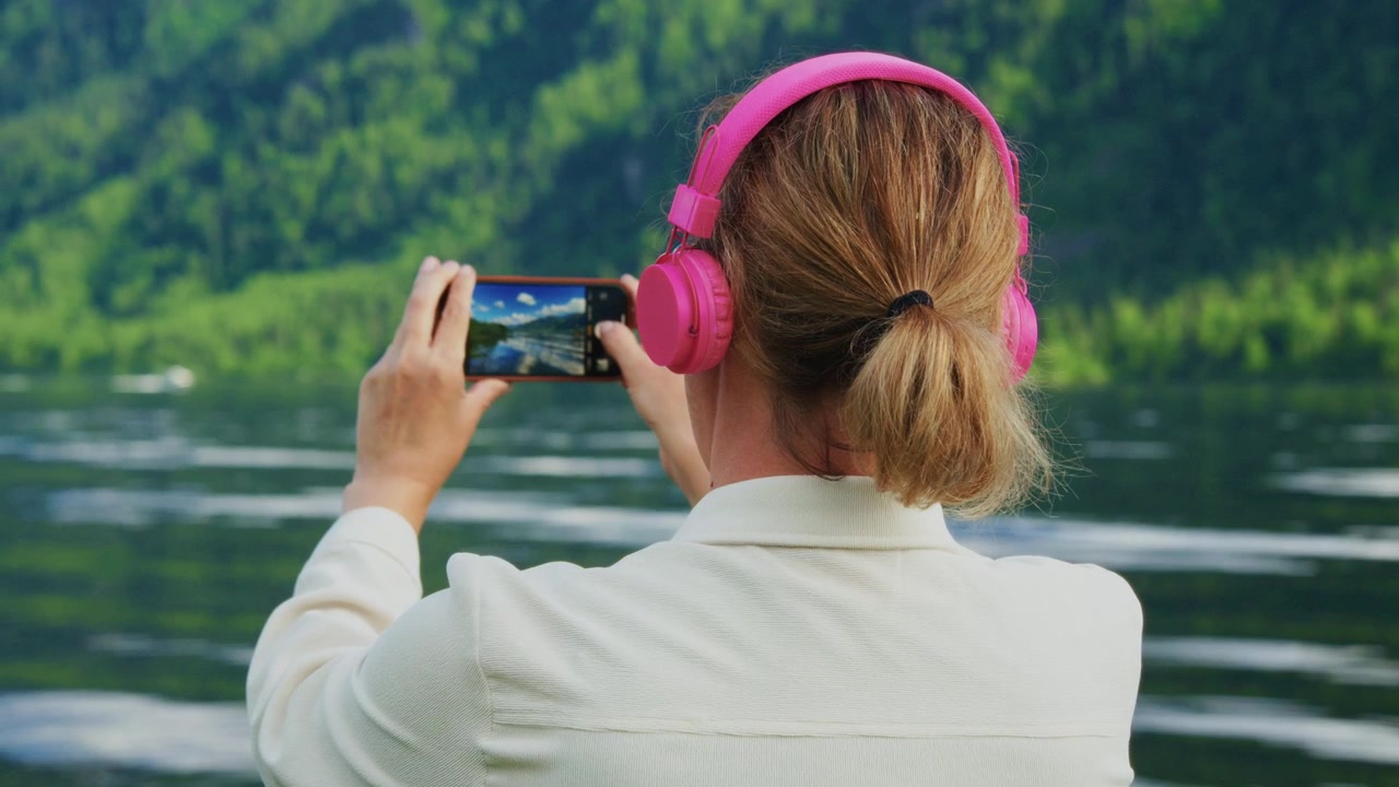 Una donna indossa cuffie rosa e scatta foto in riva al lago. È calma, concentrata a catturare momenti panoramici. Montagne verdi vibranti circondano la superficie tranquilla dell'acqua. Ideale per viaggi, relax
