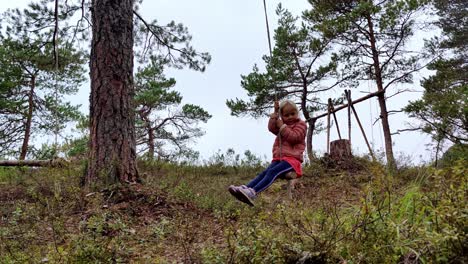 Young girl in pink riding natural swing made of rope and a branch - Having fun outdoors alone in nordic pine forest