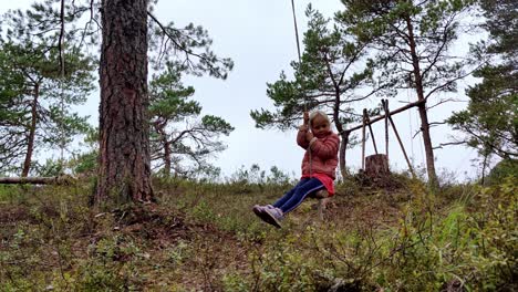 young girl in pink riding natural swing made of rope and a branch - having fun outdoors alone in nordic pine forest