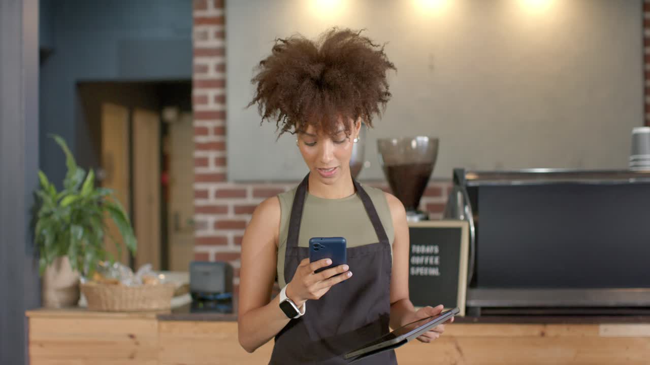 African American barista taking call and checking order on smartphone and tablet at counter