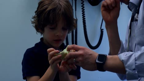 Vista-Lateral-De-Un-Joven-Médico-Asiático-Mostrando-Un-Modelo-De-Oreja-A-Un-Paciente-Niño-Caucásico-En-La-Clínica-4k