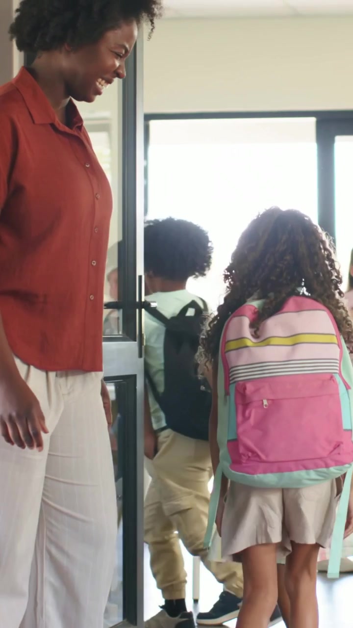 Vertical video: Smiling rust-top teacher walking in class as kids entering with packs, copy space