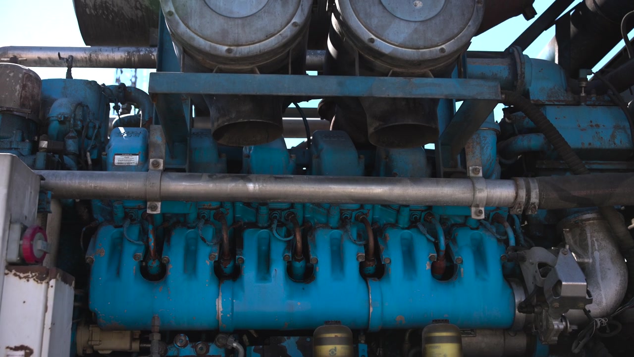 Low angle close view of teal blue industrial engine bay with cylinder bank, manifold, piping, filters, hoses, clamps, brackets, bolts, and worn metal surfaces in clean daylight, with engine, cylinder