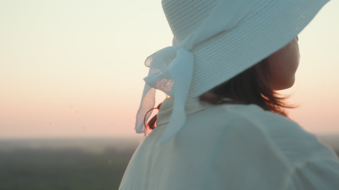 Side view of woman in white sun hat blowing soap bubble toward setting sun over river valley, her profile lit by golden glow against grassy hillside